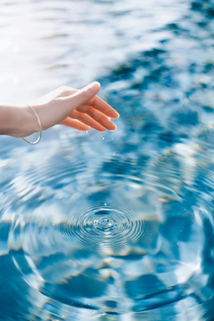 pexels-photo-15469080-15469080 A woman's hand gently creates ripples on the surface of a blue swimming pool under bright sunlight.